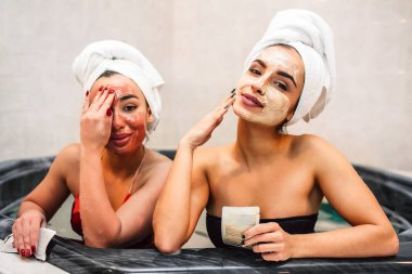 Rahat genç kadın kameraya bak. Yüzlerini beyaz ve kırmızı maske ile kaplı. Modelleri Jakuzi su dolu otur. Kadınlar Yüz Çamur maskesi üzerine dokunma.