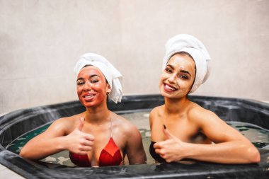 İki genç modeller büyük başparmak tutun ve doğrudan kameraya bak. Onlar gülümseme. Genç kadınların güzellik maskeleri yüz ve saç çevresinde havlu var. Modelleri jakuzide oturun.