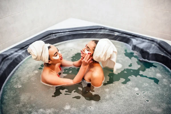 Dwie seksowne piękne młode kobiety siedzą razem w jacuzzi. Azjatycka modelu umieścić niektóre plamy oczu na twarzy europejskich przyjaciół. Spędzają czas w wodzie z pianki. — Zdjęcie stockowe