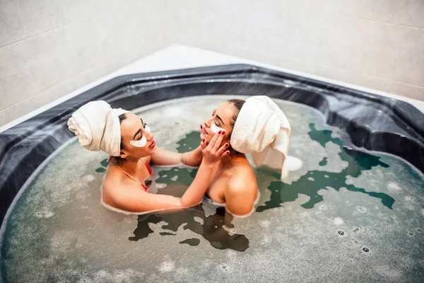 Duas jovens sensuais e encantadoras sentam-se juntas em jacuzzi. Modelo asiático colocar alguns remendos de olho em seu rosto amigos europeus. Eles passam tempo na água com espuma . — Fotografia de Stock