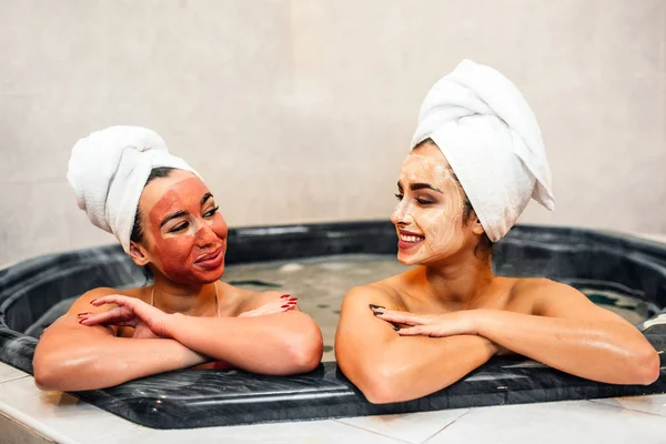 Delightful and cheerful young women relaxing in jacuzzi. Theu keep hands crossed and look at each other. Models smile. They have mask on face and towels on hair.