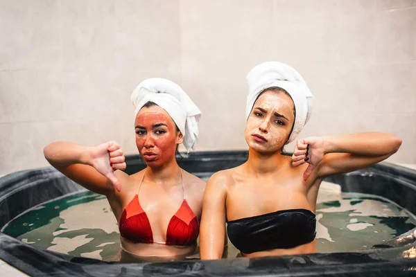 Üzgün genç kadın jakuzi ve su ile otur. Onlar mutsuz. Modelleri büyük başparmak basılı tutun. Genç kadınlar var yüz maskesi, mayo ve havlu arounf saç.