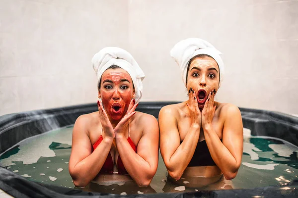 Sorprendida mujeres jóvenes se sientan en el jacuzzi y hacer procedimientos de belleza. Tienen frío y relajarse. Los modelos parecen preguntados. Tocan la cara con las manos . —  Fotos de Stock