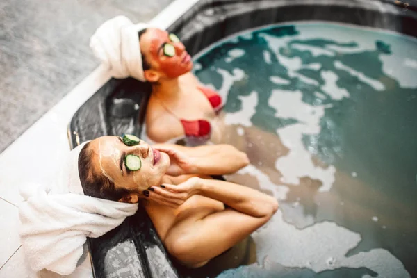 Las mujeres jóvenes relajadas tienen procedimientos de spa en el jacuzzi. Tienen mascarilla facial y trozos de pepino encima de los ojos. Los modelos se ven cómodos y a trozos . —  Fotos de Stock