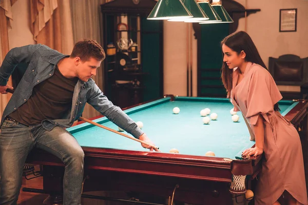 Young man hold bridge position to break ball. He is concentrated. Young woman in pink dress looks at him. She smiles. They alone in playing room. — Stock Photo, Image