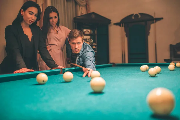 Two beautiful models watch how redhead man plying billiard. He stand in angled position with billiard cue and aiming to break ball. They are in room. — Stock Photo, Image