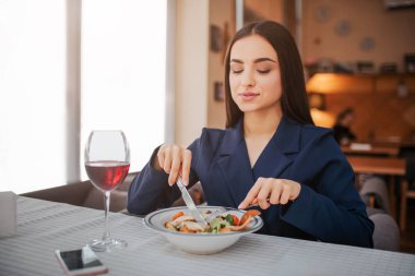 Proud and nice young woman sit at table and eat salad with fork and knife. She look down. Glass of red wine stand besides. Phone lying on table clipart