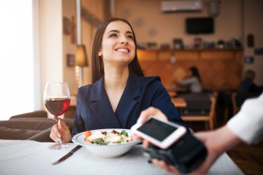 Neşeli genç müşteri masasına oturup yukarı bak. O gülümsüyor. Model telefon terminal ödeme üzerine tutun. O başka bir elinde kadeh kırmızı şarap vardır