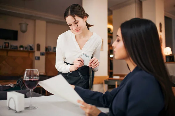 Young waitress stand besides sitting customer and look at menu she point. Businesswoman talking to her. They are in restaurant. — Stock Photo, Image