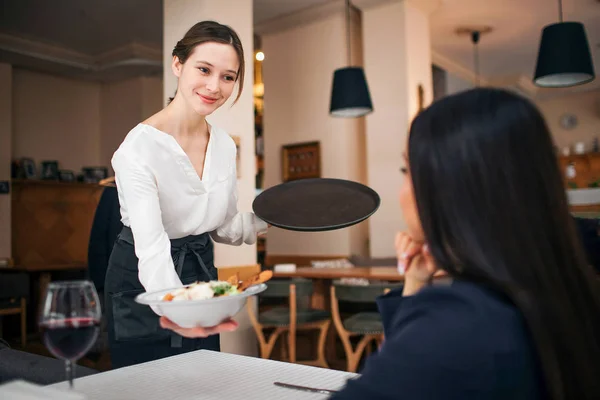Hoş genç garson masada oturan müşteri ile stand ve onu kase salata göster. O gülümsüyor. Kadın müşteri bak. Restoranda olduklarını. — Stok fotoğraf