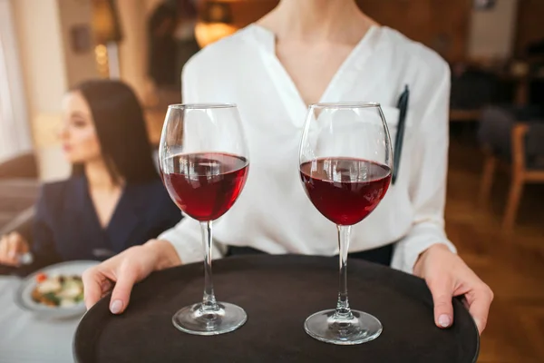 Skär vy av servitris hålla två glas rött vin på bricka. Youn affärskvinna sitta bakom henne vid bordet och titta vänster. Hon har Salladsskål på bord. — Stockfoto