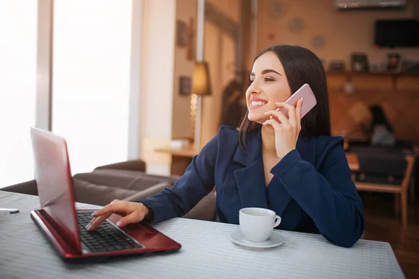 Glad ung kvinna sitter vid bord och leende. Hon prata telefon och typ på tangentbordet samtidigt. Modellen har kopp kaffe på bordet. — Stockfoto