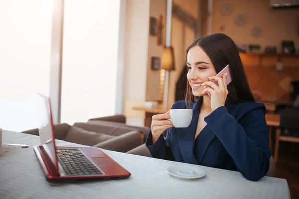 Glad ung kvinna sitter vid bord och arbete. Hon ler. Kunden prata telefon och titta på laptops skärm. Ung kvinna dricka kaffe från cup. — Stockfoto