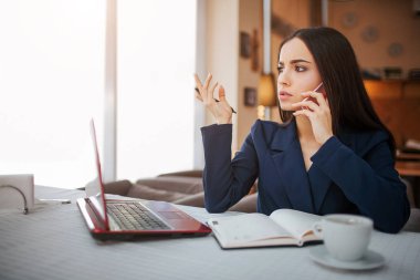 Düşünceli genç kadın masaya oturmak ve bekliyoruz. Telefonla konuşuyor. İşkadını dizüstü fincan kahve ve masa üstünde defter vardır.