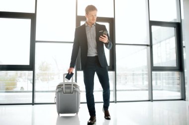 Ciddi genç işadamı iletmek için Havaalanı penceresinden yürümek. Suitcaase rulo ve siyah telefon elinde tut. Adam göz