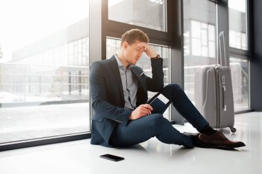 Genç adam baş ağrısı vardır. Havaalanı salonu penceresinde, katta oturmak. Adam bilet pasaport ile elinde tut. Telefon ve bavul yanında bulunmaktadır.