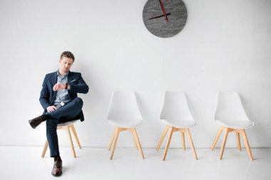 Genç adam oturup beyaz sandalye ve uçuş için bekleyin. Saatler baktığını. Adam içinde pasaport ve bilet tutun. O tek başına odasında oturuyor.
