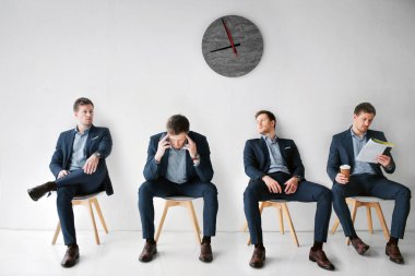 Concept waiting for a meeting. Four young man in suits sitting on white chairs in one room. First look to right. Second has headache. Third lean to wall. Fourth read menu and hold cup of coffee. clipart