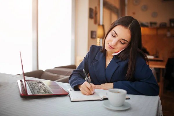 Trevlig ung kvinna sitta vid bord och prata på telefon. Hon skriver i anteckningsboken. Affärskvinna arbete i restaurang. Hon har laptop på tabell. — Stockfoto