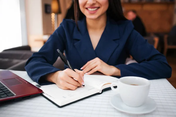 Skär syn på glad ung kvinna leende och skriva i anteckningsbok. Hon har laptop och kopp kaffe vid bordet. Ung affärskvinna arbete i restaurang. — Stockfoto