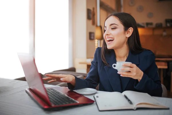 Aufgeregte und gut gelaunte junge Frau sitzt am Tisch im Restaurant. Sie zeigt auf den Bildschirm des Laptops. Modell lächelnd. Sie hat Notizbuch mit Stift auf dem Tisch. — Stockfoto