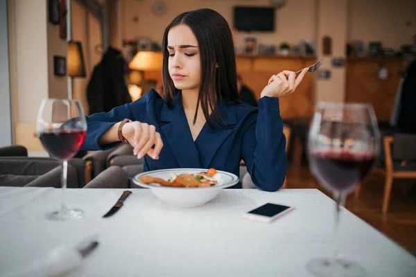 Несчастная молодая женщина сидит за столом и обедает. Она смотрит на часы. Модель раздражена. Два бокала красного вина стоят за столом . — стоковое фото