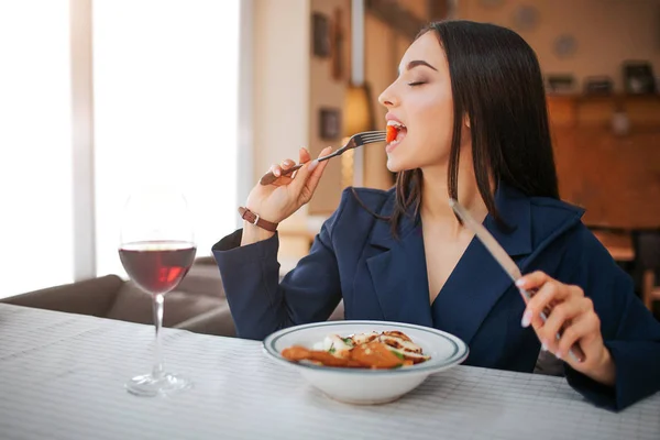 Ung kvinna sitter vid bordet i restaurangen och njuta av att äta sallad. Hon hålla ögonen stängda. Glas rött vin står på bordet. — Stockfoto