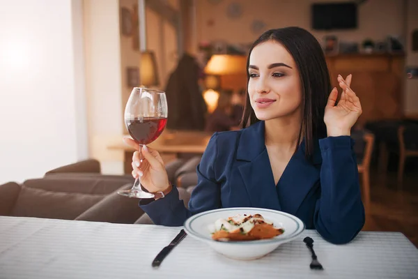 Vacker ung affärskvinna sitta vid bordet i restaurangen. Hon ser till vänster. Kunden håller glas rödvin i handen. Salladsskål stå på bordet. — Stockfoto