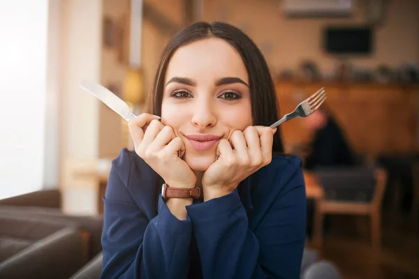 Гарна молода жінка виглядає ближче і прямо на камеру. Вона тримає ніж і виделку в руках. Модель сидить за столом у ресторані . — стокове фото