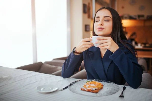 Ung kvinna njuter av sin kopp kaffe. Hon sitter vid bord och lukta på den. Modellen har kaka på tallrik på bordet. — Stockfoto