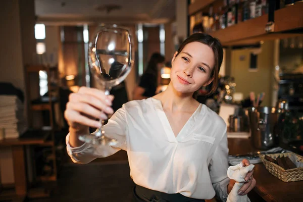 Genç garson resmini stand ve temiz bardak şarap için bak. Başka bir elinde beyaz havlu tutun. Garson iş adlı bar-restoranda. — Stok fotoğraf