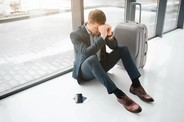 Worried and tired young man sit on floor in airport. He hide head with hands. Suitcase and phone with tickets are besides. — Stock Photo, Image