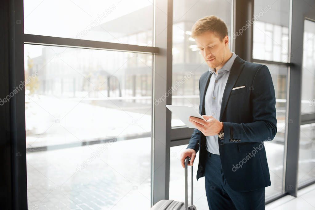 Handsome young businessman stand in airport hall. He hold hand of suitcase and white tablet. Guy smiles. He stand at window.