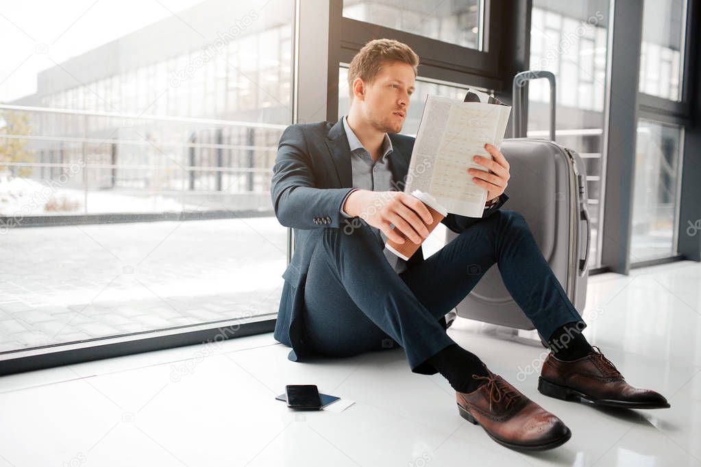 Hungry young man sit on floor and wait for flight. He hold menu and look at it. Also guy has cup of hot drink. He left phone and suitcase on floor besides.