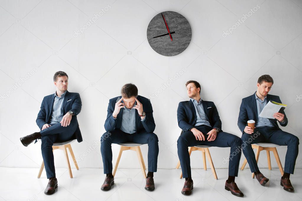 Concept waiting for a meeting. Four young man in suits sitting on white chairs in one room. First look to right. Second has headache. Third lean to wall. Fourth read menu and hold cup of coffee.