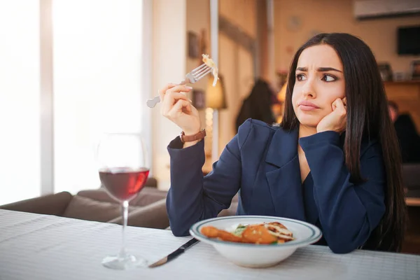 Zmatená mladá žena sedí u stolu a podívat se na kousek salátu ona drží. Její zrak je divný. Youhng žena sedět v restauraci. Salát nad sklenicí vína jsou na stole. — Stock fotografie