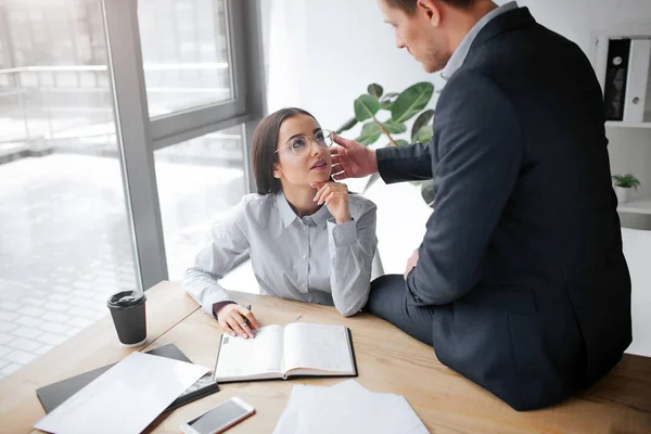 Pictrue íntima de mujer joven encantadora sentarse en la mesa y mirar a su jefe. Se sienta en la mesa y le toca la mejilla. El tipo está sexualmente excitado. Están en una habitación . — Foto de Stock