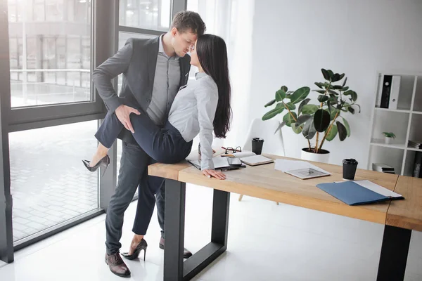 Pár munkahelyi szexuális és hangulatos kép. Ő ül az asztalon. Ő tartsa a lábát a szexuális jelent. Ők eltartás arcok egymáshoz közel. — Stock Fotó