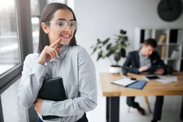 Glad och positiv ung kvinna stå och leende. Hon trycker du på håret och se tillbaka på boss. Han sitter vid bordet. Guy titt på brunett. — Stockfoto