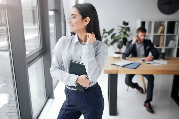 Glad ung kvinna stå och pose. Hon ler och titta på fönstret. Hennes chef är upptagen. Han prata på telefon. De är i ett kontorsrum. — Stockfoto