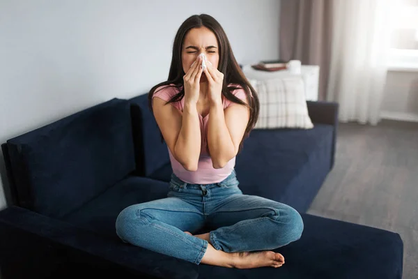 Photo d'une jeune femme assise sur un canapé et éternuant dans une serviette blanche. Elle est malade. Le mannequin souffre. Elle se sent mal. . — Photo