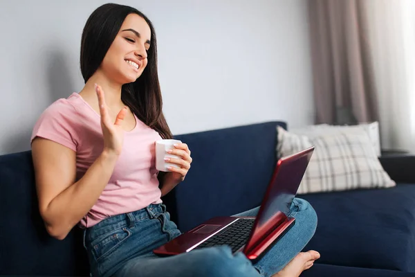 陽気な若い女性は、ソファと手で波の上に座る。彼女は微笑んで、ノート パソコンの画面を見ています。モデルは、nap にそれを保持します。彼女は飲み物の白いカップ. — ストック写真