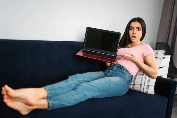 Naštvaná a roztržitý mladá žena sedět na pohovce v pokoji. Ona drží červené laptop. Mladá žena bod na černé obrazovce. Notebook je přerušeno. Nefunguje to. — Stock fotografie