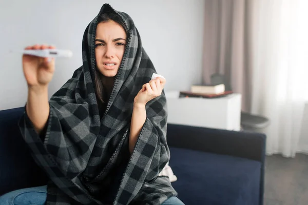 Una joven enferma sostiene el termómetro en la mano. Se lo mostró a la cámara. El modelo tiene temperatura. Se cubre con una manta. . — Foto de Stock