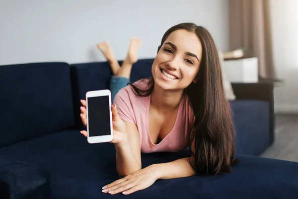 Jovencita alegre acostada boca abajo en el sofá. Ella sostiene el teléfono blanco y sonríe a la cámara. Modelo mostrar dispositivo con pantalla oscura . — Foto de Stock