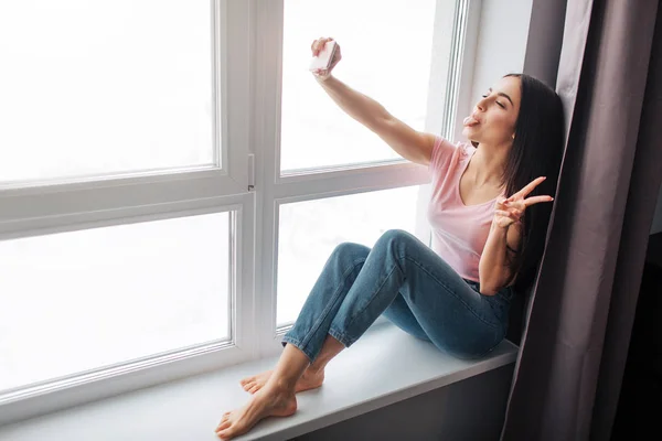 Jovencita alegre tomar selfie en la ventana. Sonríe y posa. Modelo espectáculo pieza signo con los dedos . —  Fotos de Stock