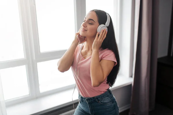 Une jeune femme calme et gentille se tient dans la chambre. Elle aime écouter de la musique. Modèle garder les yeux fermés et les mains sur les écouteurs . — Photo