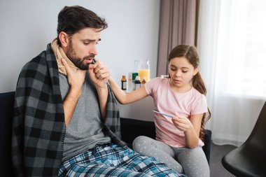 Hasta genç adam oturup kanepe ve öksürük. Yumruk ediyor. Boğaz eşarp ile kaplıdır. Omuzlarında yalan battaniye. Kız termometre bak ve elini onun omzuna tut.