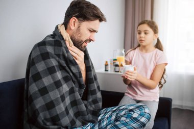 Hasta genç adam oturmak ve battaniyeye örtülü. O el ele tutuşsun boğaz üzerinde. Ciddi küçük kız ona bakmak ve beyaz Kupası tutun.