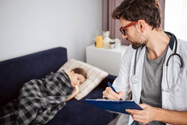 Hasta bir küçük kız yatakta yatarken battaniye ile örtülü. Genç erkek doktor ona bak ve yaz. Boynunda asılı stetoskop.